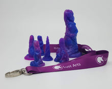 Load image into Gallery viewer, Photo of a medium-dark purple lanyard with the Lust Arts logo in white with a silver hook on an off-white background with a Mermaid adult toy and a few charm versions
